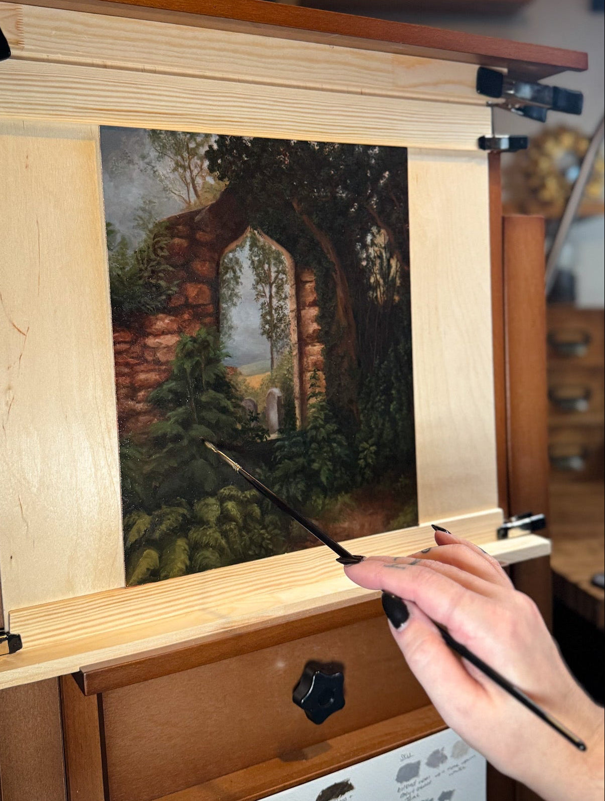 Laura Miller painting a Scottish church ruin landscape scene in oil paint on a wooden easel with a brush.