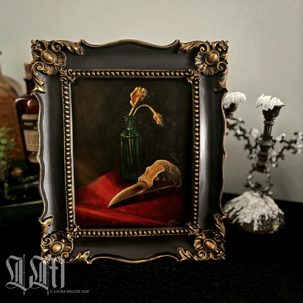 Decorative still life painting in a baroque style, with a crow skull and dried yellow flowers in vintage green bottle. Framed in ornate black and gold antique style picture frame.